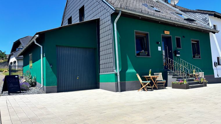 Ein modernes, grünes Haus mit einer Eingangstreppe und einem kleinen Terrassenbereich. Vor dem Haus befinden sich gepflasterte Flächen und blühende Pflanzen.