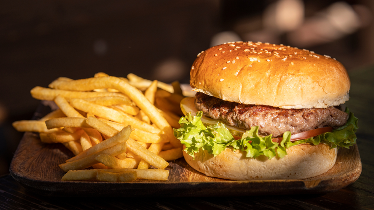 A juicy burger with a sesame bun, lettuce, and tomato is placed on a wooden board. Next to it are crispy French fries.