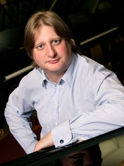 Ein Mann sitzt am Klavier und schaut in die Kamera. Er trägt ein hellblaues Hemd und hat blonde Haare.
