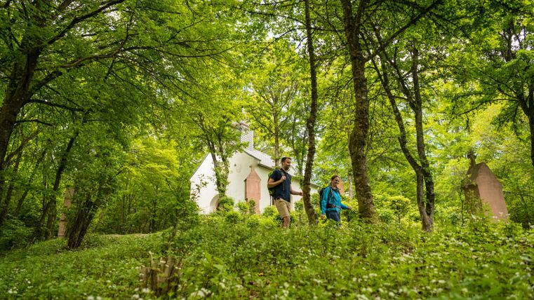 Zwei Wanderer gehen durch einen Wald mit einer kleinen weißen Kapelle im Hintergrund.