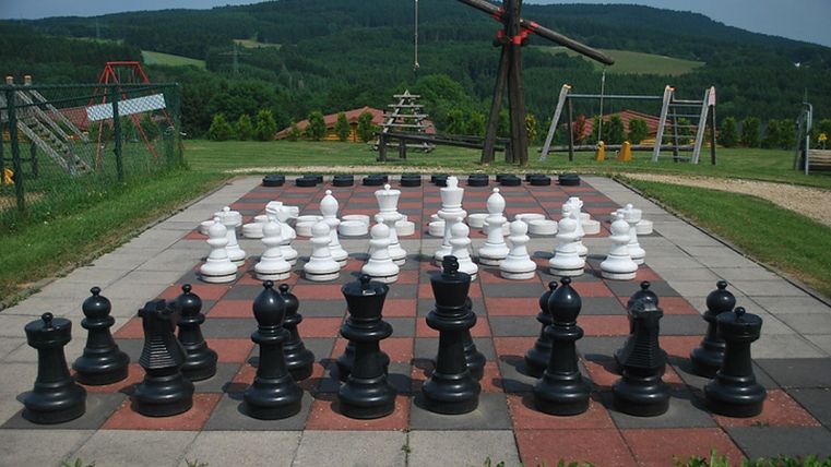 Großes Schachbrett im Freien mit schwarzen und weißen Figuren. Im Hintergrund sind grüne Hügel und Spielplatzgeräte zu sehen.