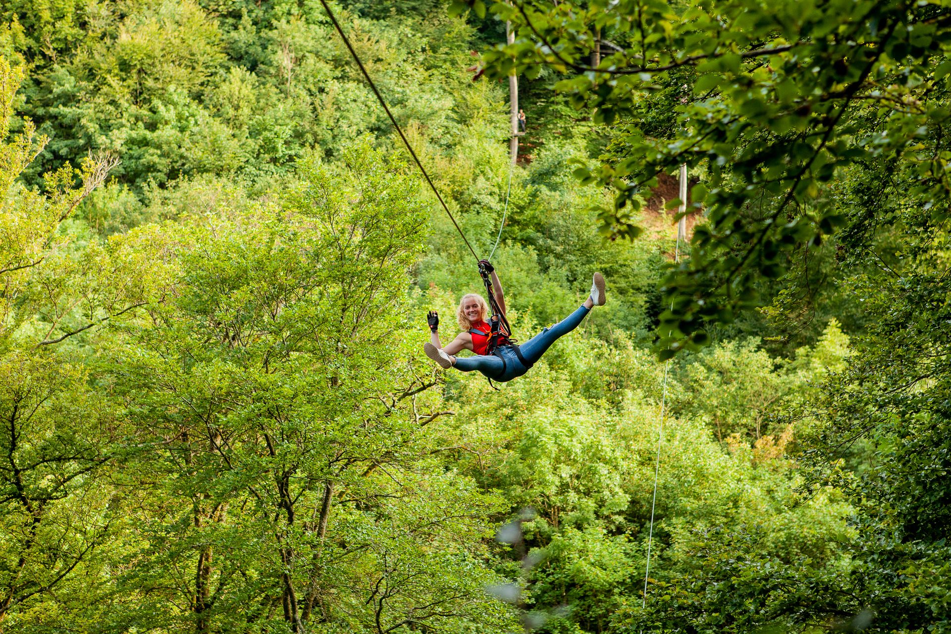Eine Frau ist mit Gurten und Sicherungsgeschirr an eine Zipline geschnallt und fährt durch einen Wald.