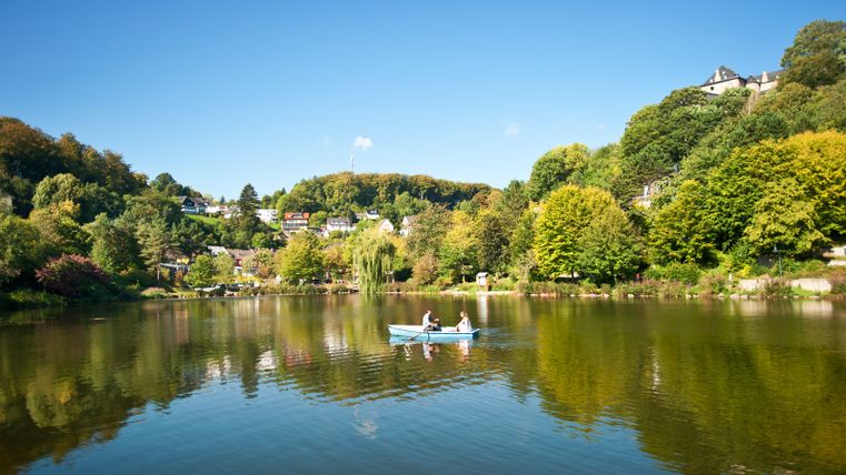 Un étang à Blankenheim avec un bateau à rames et deux personnes, entouré d'arbres et de maisons sur une colline.