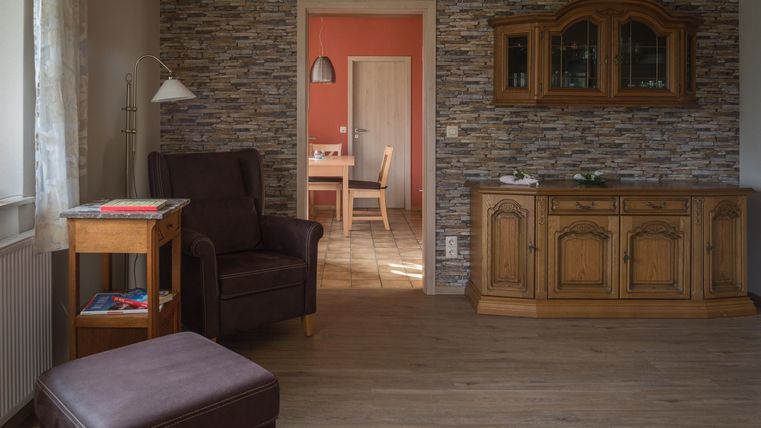 A cozy living space with a brown armchair and a wooden sideboard. In the background, another room with a dining table is visible.