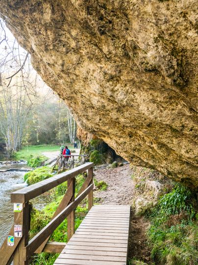 Blick unter einem massiven Felsen hindurch zu einer Holzbrücke, die über einen Bach nahe des Felsens führt.