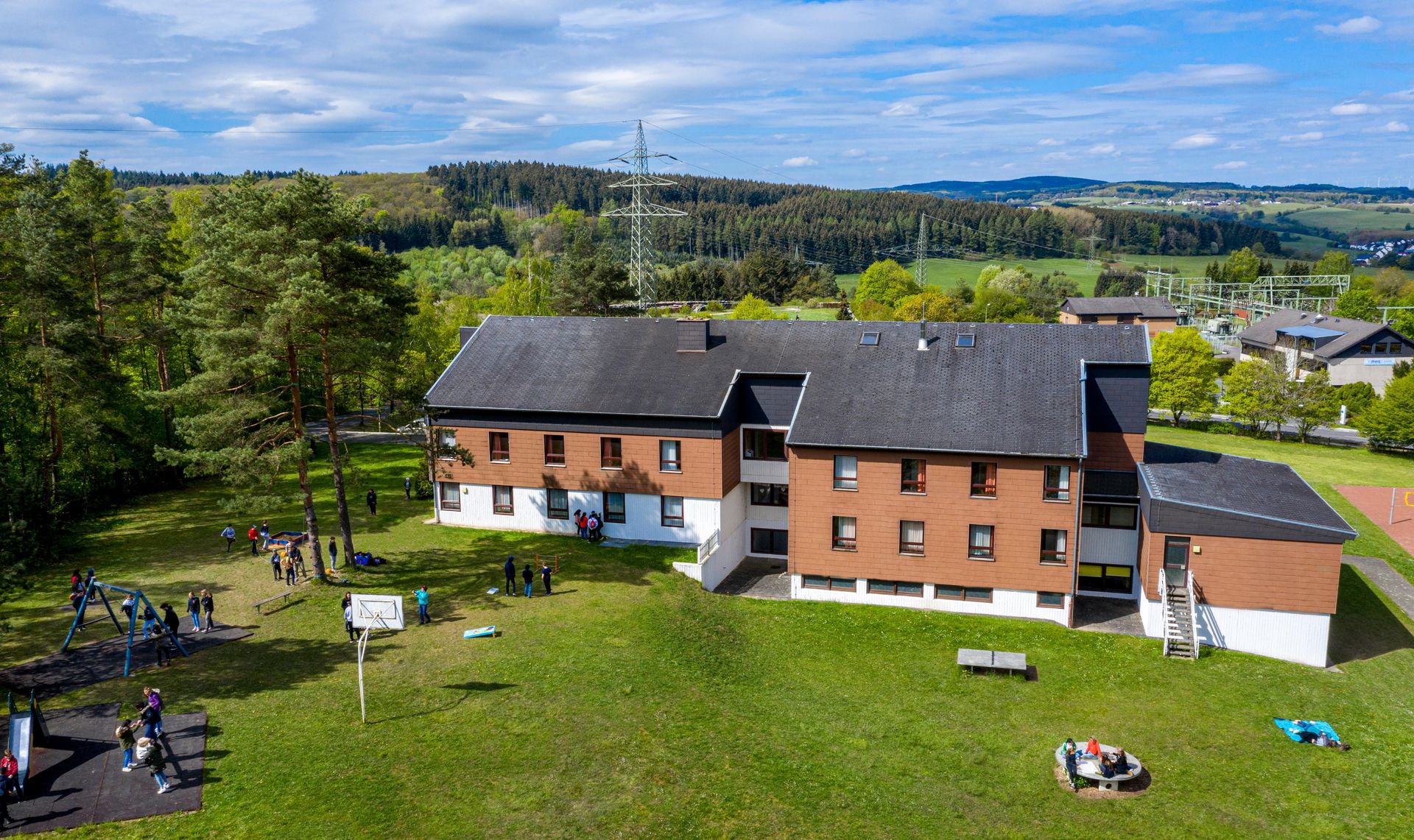 Eine große Jugendherberge liegt auf einer Anhöhe auf einer Wiese am Rande eines Waldes. Besucher verteilen sich rund um das Gebäude.