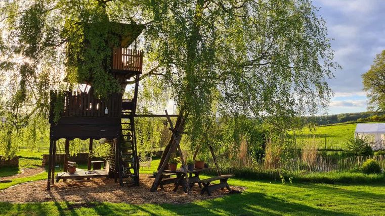 Une tour de jeu en bois sous un grand arbre, entourée d'herbe verte et d'un petit étang. Le soleil brille et projette des ombres sur la pelouse.