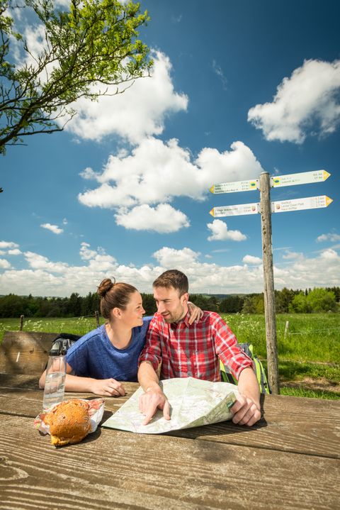 Ein Mann und eine Frau sitzen auf einer Rastbank inmitten der Natur und studieren eine Wanderkarte auf dem Tisch.