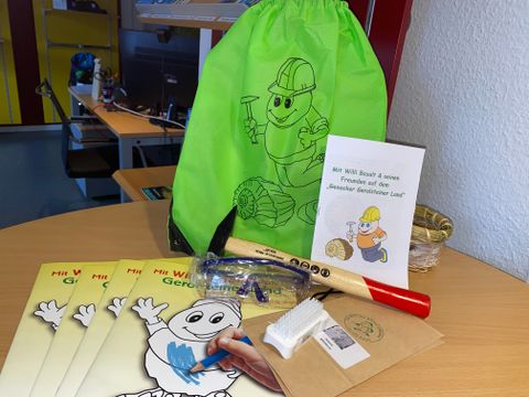 Auf einem runden Holztisch liegt die Forscherausrüstung für den Gerolsteiner Geoacker: ein grüner Stoffbeutel, mehrere Malhefte, ein Hammer, eine Schutzbrille, eine Bürste und ein Infoheft. 