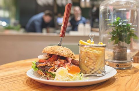 Auf einem Holztisch steht ein Teller mit einem Burger und Pommes darauf, in dem Burger steckt ein Messer, im Hintergrund sieht man arbeitende Menschen