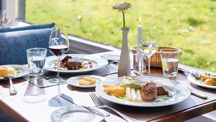 Elegantes Mittagessen mit Fleisch, Spargel, Rot- und Weißwein auf einem gedeckten Tisch am Fenster mit Blick ins Grüne.