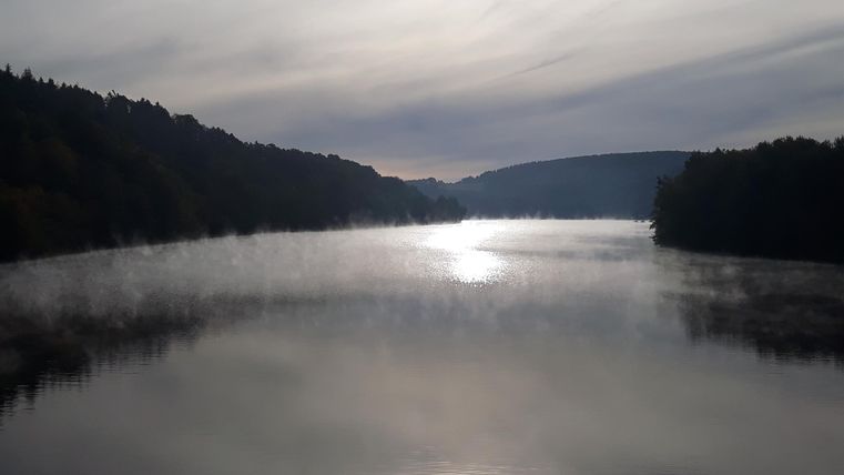Ein ruhiger See mit sanften Nebelschwaden und den umliegenden Bergen. Die Spiegelung des Lichts auf dem Wasser schafft eine friedliche Atmosphäre.