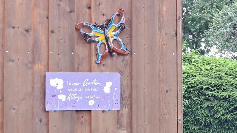 Ein schöner Schmetterling aus buntem Material hängt an einer Holzwand. Darunter befindet sich ein Schild mit einer einladenden Botschaft.