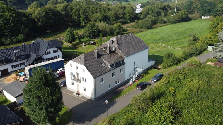 Luftaufnahme eines Gebäudekomplexes in ländlicher Umgebung, umgeben von Bäumen und Wiesen. Mehrere Autos sind auf dem Parkplatz zu sehen.