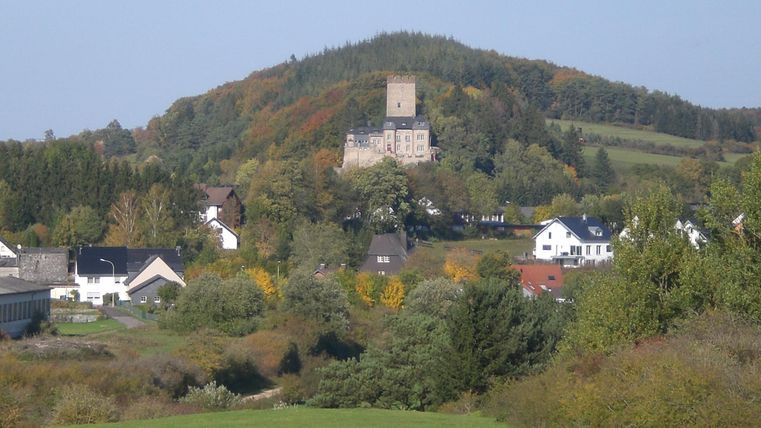 Eine malerische Landschaft mit sanften Hügeln und einem Schloss auf einem Berg. Im Vordergrund sind grüne Wiesen und einige Häuser zu sehen.