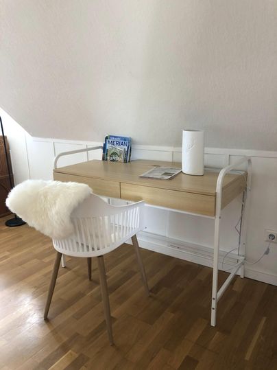 Ein moderner Schreibtisch mit einem weißen Stuhl steht in einem hellen Raum. Auf dem Tisch liegen einige Zeitschriften und eine Lampe.