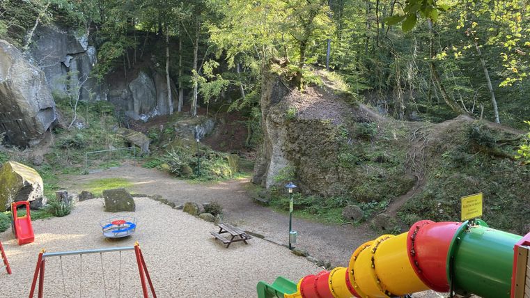 Spielplatz mit bunter Rutsche, Schaukel und Klettergerüst in einer felsigen, bewaldeten Umgebung.