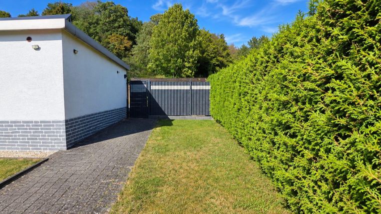 Un chemin de jardin bien entretenu à côté d'une haute haie verte. En arrière-plan, on voit un bâtiment clair et un ciel bleu.