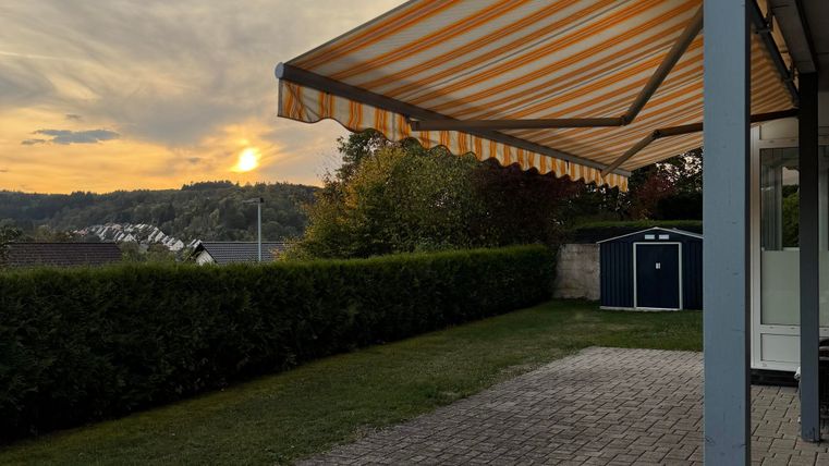 Un jardin avec un parasol et une belle ambiance du soir. À l'arrière-plan, le soleil couchant et des collines douces sont visibles.