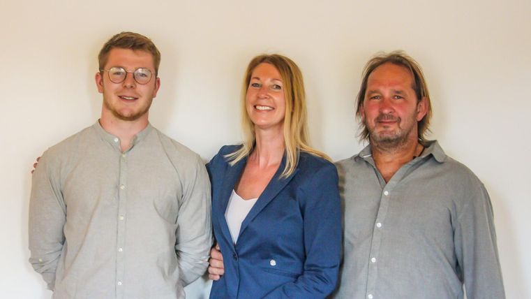 Drei Personen stehen zusammen vor einer neutralen Wand. Sie lächeln und wirken freundlich.