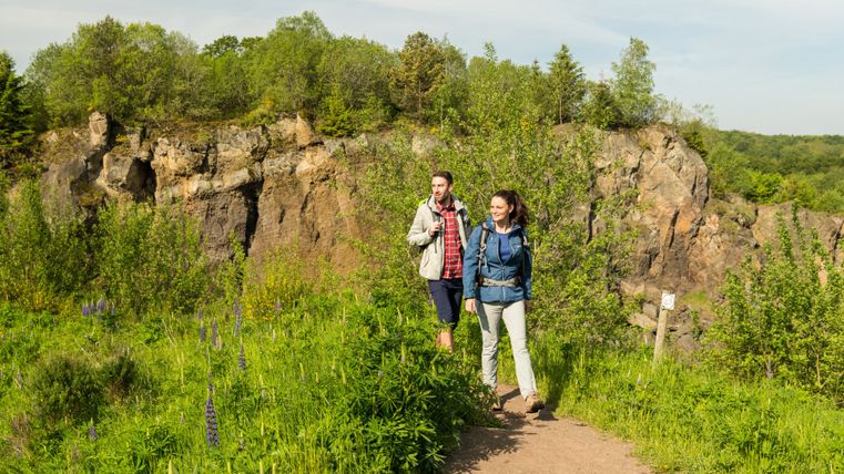 Zwei Personen wandern auf einem Pfad durch eine grüne Landschaft mit Felsen im Hintergrund.