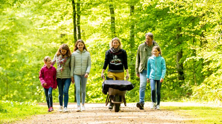 Eine Familie spaziert durch einen Wald und eine Frau in der Mitte schiebt eine Schubkarre vor sich.