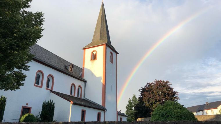 Eine Kirche mit einem spitzen Turm steht im Vordergrund. Im Hintergrund sieht man einen Regenbogen und eine grüne Landschaft.