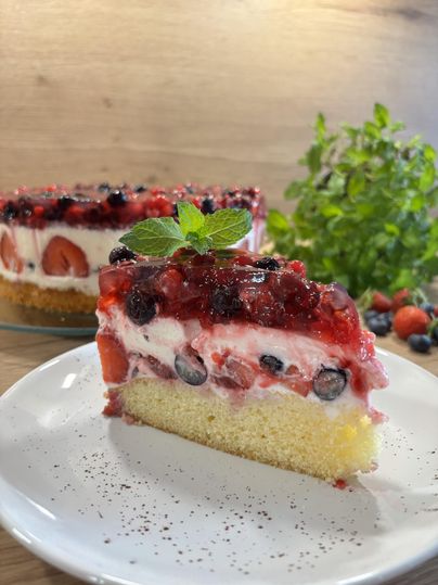 Ein Stück Waldfruchttorte bestehend aus einer Schicht Beeren in Gelee, darunter einer weißen Creme und darunter Kuchenboden steht auf einem Teller. Im Hintergrund die restliche Torte.