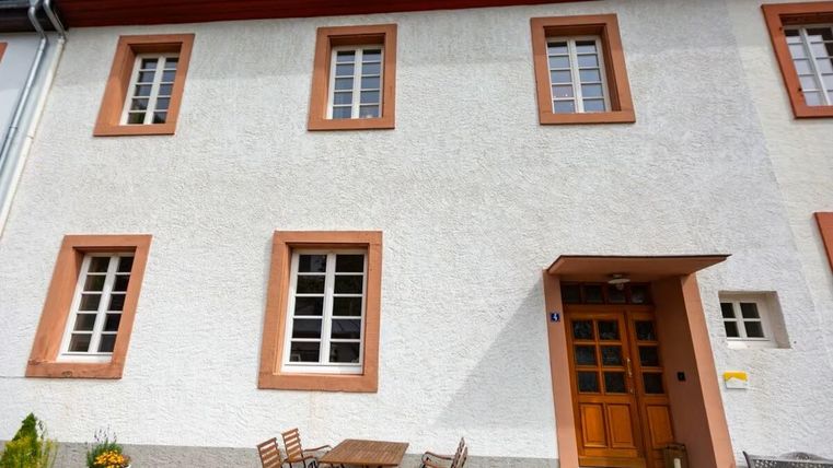 Ein helles, einladendes Gebäude mit weißen Wänden und orangefarbenen Fensterrahmen. Vor der Tür steht ein Tisch mit Stühlen, ideal für entspannte Stunden im Freien.