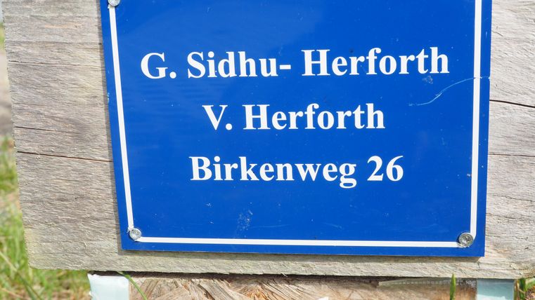 Ein blauer Schild mit dem Namen "G. Sidhu-Herforth" und "V. Herforth" steht an einem Holzpfosten. Die Adresse "Birkenweg 26" ist ebenfalls auf dem Schild vermerkt.