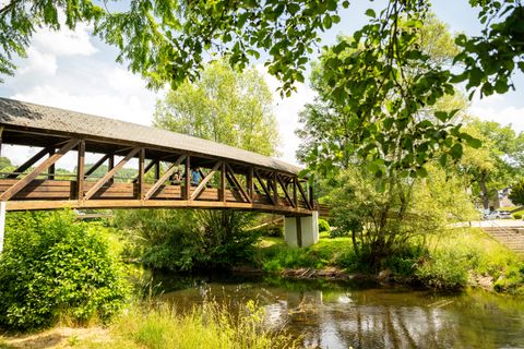 Eine überdachte Holzbrücke führt über einen Fluss in einem reichlich bewachsenen Parkgelände.