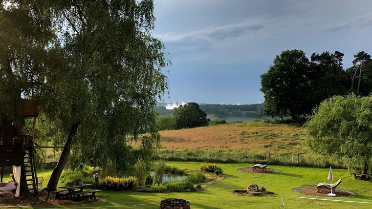 Une pelouse verte avec des arbres et un petit étang. À l'arrière-plan, on peut voir des collines et un ciel nuageux.