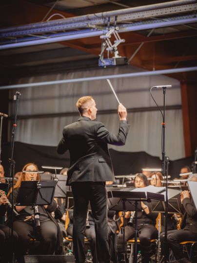 Ein schwarz gekleideter Dirigent vor einer großen Gruppe Musikanten mit unterschiedlichen Instrumenten.