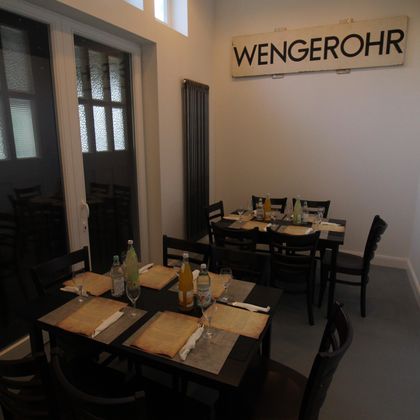 Ein Restaurant mit dunklen Holztischen, gedeckt mit Gläsern und Getränken. An der Wand hängt ein Schild mit der Aufschrift 'WENGEROHR'.