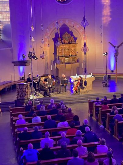 Fünf Musiker an Streichinstrumenten und Klavier musizieren vor dem Kirchenaltar. Davor die Sitzbänke voller Besucher.