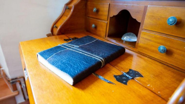 Ein schwarzes Notizbuch liegt auf einem hölzernen Schreibtisch. Der Schreibtisch hat eine schöne, warme Holzmaserung und einige kleine Schubladen.