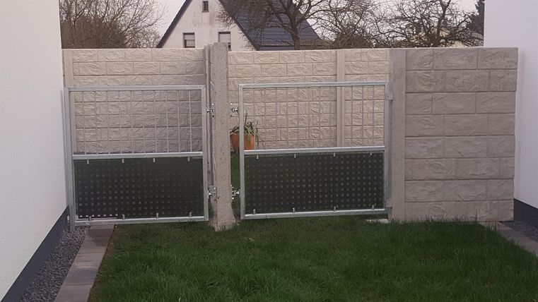 Ein Eingang mit zwei metallenen Toren und einer Steinmauer. Im Hintergrund sind Bäume und ein Haus sichtbar.
