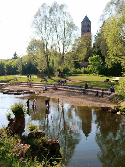 Menschen genießen einen sonnigen Tag im Kyllpark mit einem Kirchturm im Hintergrund.