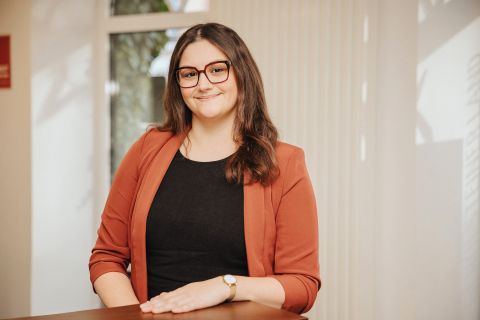 Eine Frau steht hinter einem Tresen in einem hellen Büroraum und lächelt in die Kamera. Hinter ihr einige Fenster.