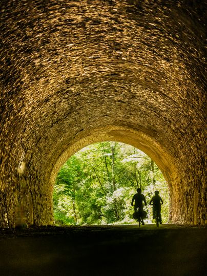 Zwei Radfahrer fahren durch einen beleuchteten Tunnel mit Blick auf grüne Bäume am Ausgang.