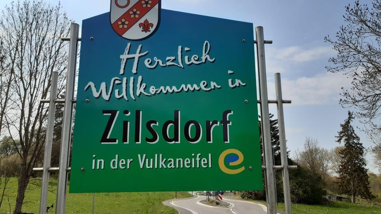 Ein Willkommensschild für Zilsdorf in der Vulkaneifel. Die Umgebung ist grün und bietet eine ländliche Kulisse.