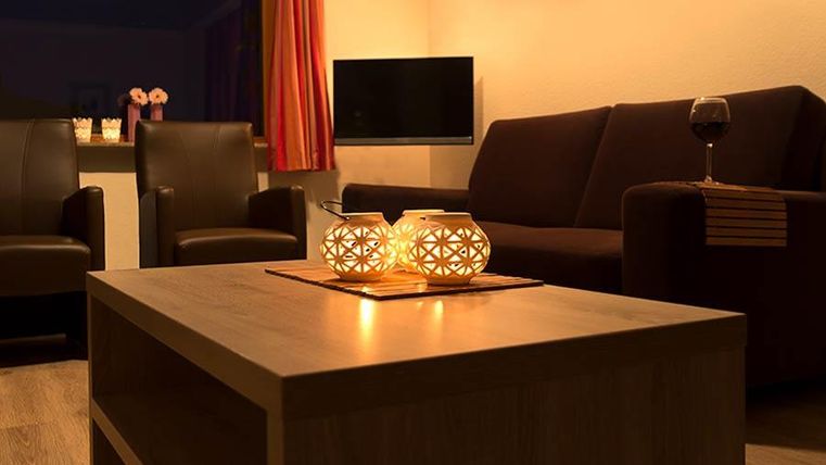 Ein gemütliches Wohnzimmer mit einem dunklen Sofa und zwei Sesseln. Der Tisch hat eine schöne Dekoration aus Kerzen und es gibt ein Glas Wein.