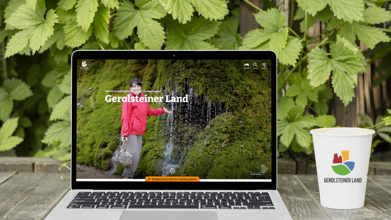 Ein Holztisch draußen vor einer bewachsenen Wand. Auf dem Tisch steht ein Laptop, auf dem die Homepage der Ferienregion Gerolsteiner Land geöffnet ist. Daneben ein Pappbecher mit dem Logo der Touristik GmbH Gerolsteiner Land darauf.