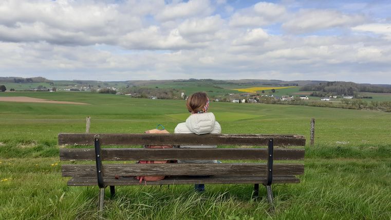 Person sitzt auf einer Bank und blickt auf eine weite, grüne Landschaft mit Feldern und Wolken am Himmel.