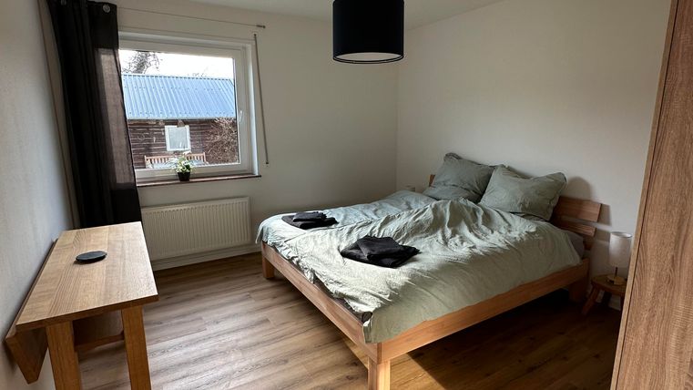 Ein einfaches Schlafzimmer mit einem Bett und einem Holzschreibtisch. Der Raum hat helle Wände und einen großen Fenster mit Tageslicht.