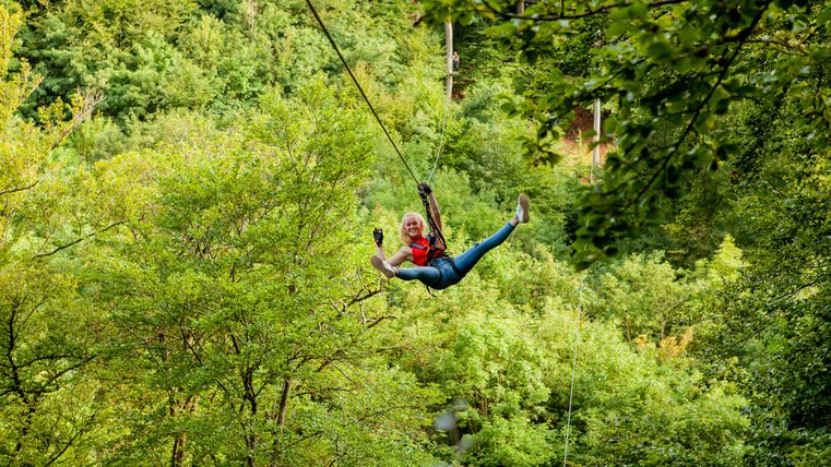 Eine Frau ist mit Gurten und Sicherungsgeschirr an eine Zipline geschnallt und fährt durch einen Wald.