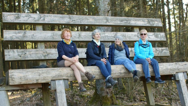 Un grand banc en bois dans la forêt, sur lequel quatre femmes sont assises. L'environnement est naturel avec des arbres en arrière-plan.