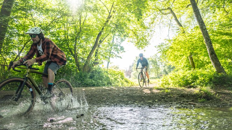 Zwei Radfahren auf Gravelbikes fahren durch eine große Pfütze mitten im Wald. Die Sonne scheint durch die Bäume hindurch.