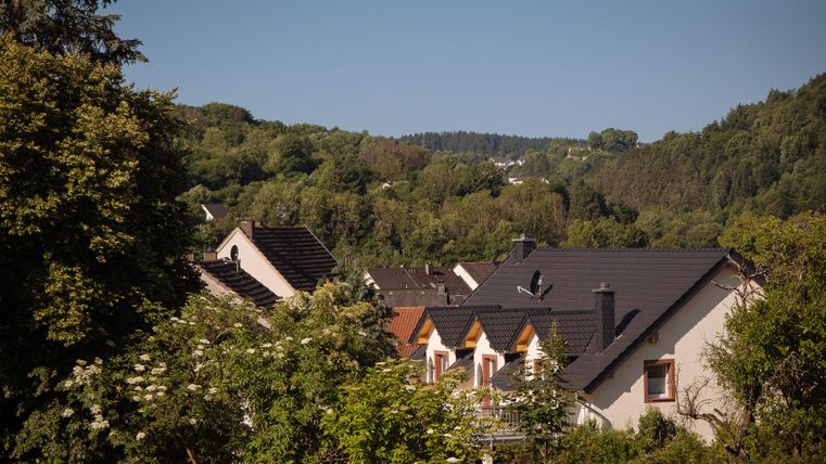 Eine ruhige Wohngegend mit schönen Häusern und viel grünem Bewuchs. Im Hintergrund sind sanfte Hügel und Wälder zu sehen.