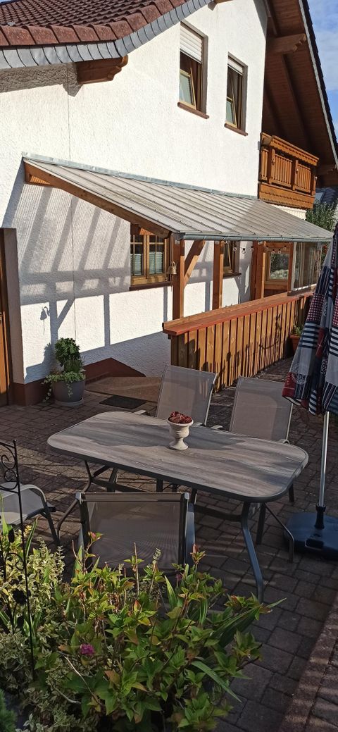 A cozy seating area outside with a table and chairs. In the background, there is a bright house and plants.